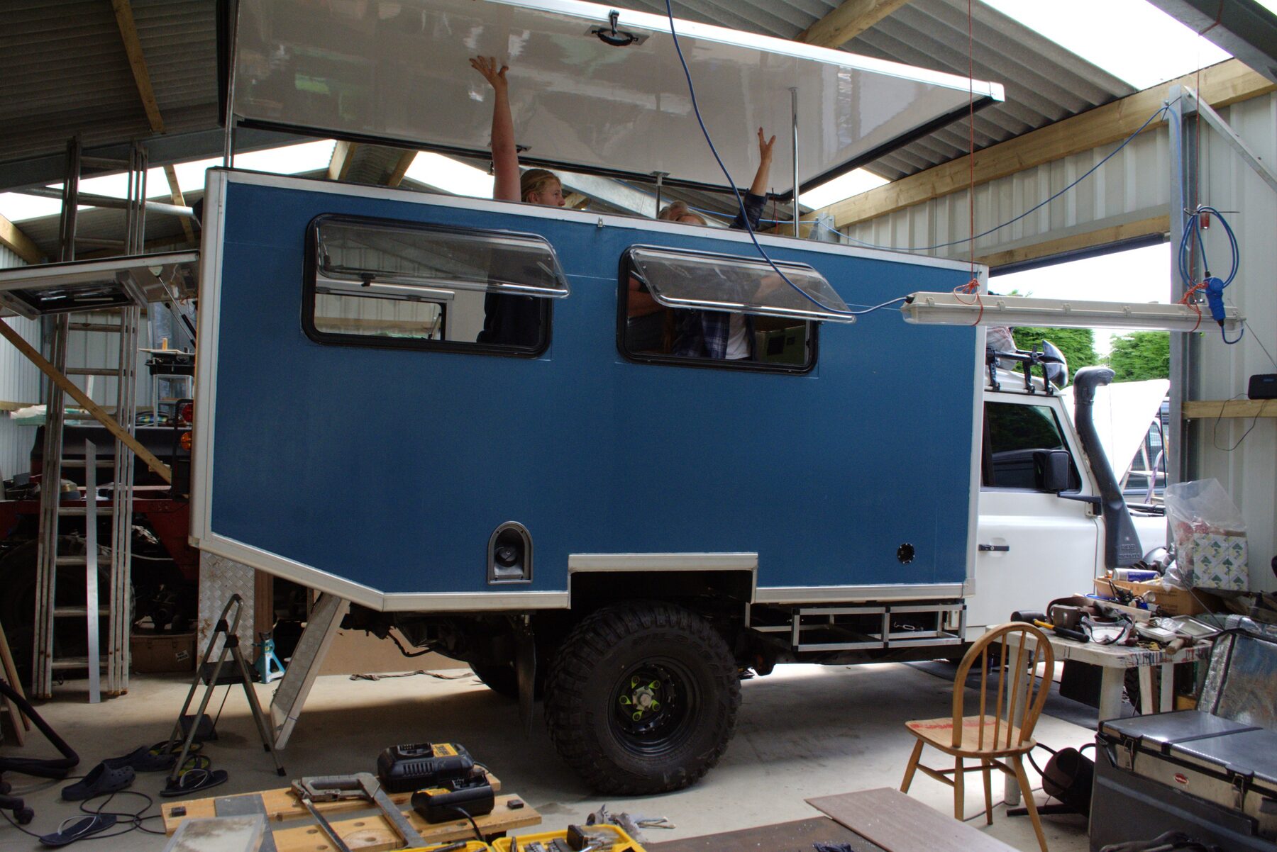 The blue camper box being assembled on the back of the Defender in a workshop, with the roof panel raised on supports and people working inside it