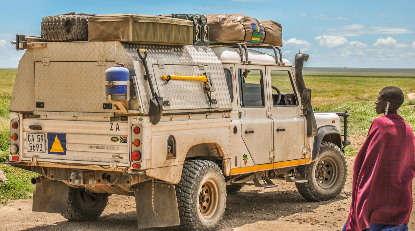 The same Defender 130 before the rebuild, seen from the rear, with an aluminium canopy, a large folded roof tent and spare wheel on the roof rack, and a person in a red cloak standing beside it