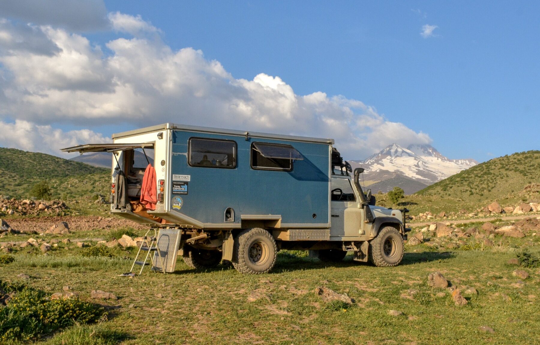 The Defender camped on a grassy hillside with a snow-capped mountain behind, the pod's roof lifted, side windows open and the large rear door swung up as an awning