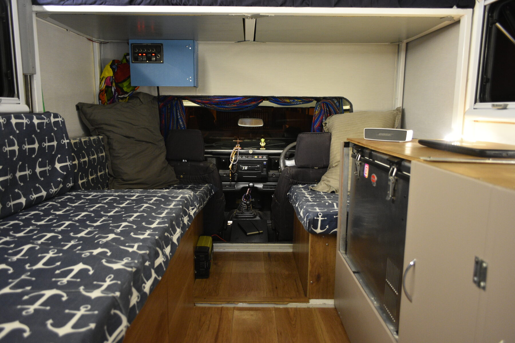Inside the pod looking forward through a wide opening to the Defender's front seats and dashboard, with patterned bench seats either side and a switch panel on the wall above the opening