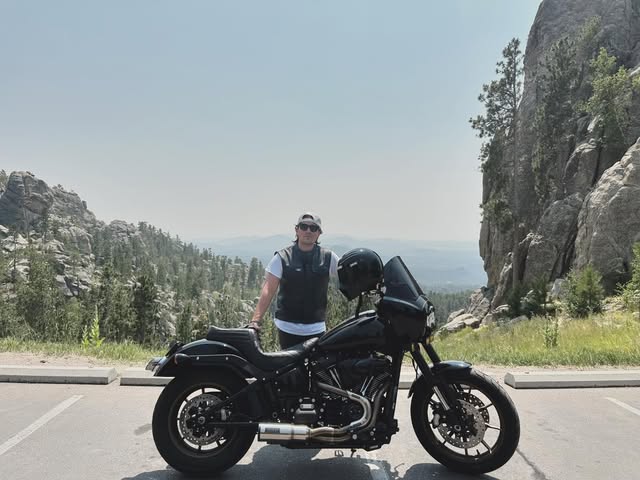 Chad Sig standing behind his black cruiser motorcycle at a pull-off on the Needles Highway, granite spires and pine forest behind him