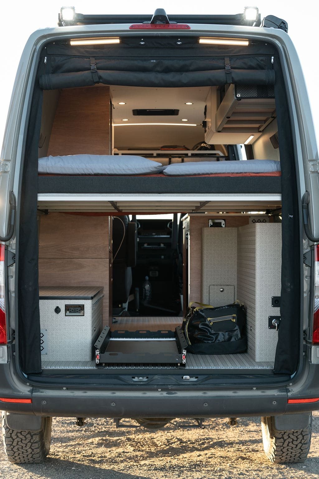 The van's rear doors open on a garage under a fixed bed platform: a low metal cabinet on the left, a tall cabinet on the right, and an empty sliding tray on the floor between them