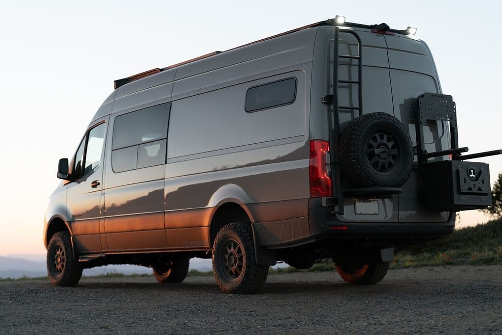 A grey high-roof Mercedes Sprinter 4x4 camper van at dusk on a gravel pullout, seen from the rear quarter, with all-terrain tires, a spare wheel and a storage box on the rear doors