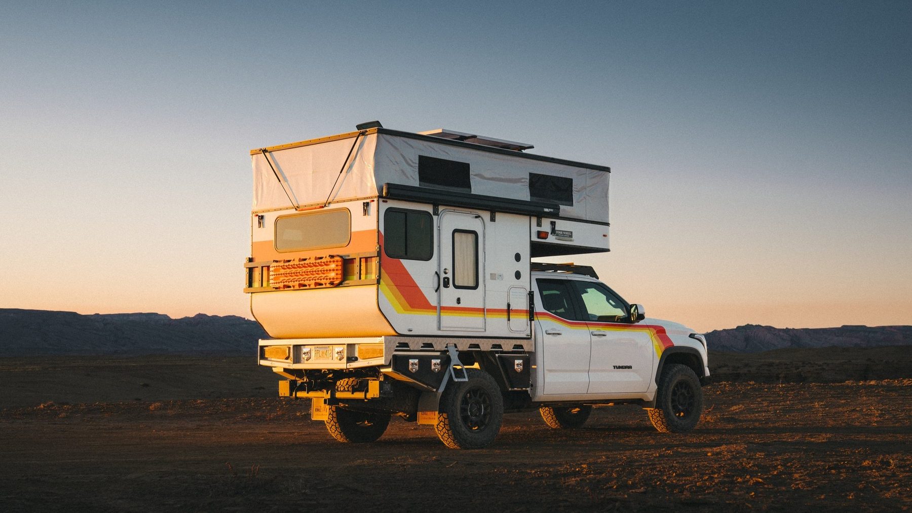 The white Tundra with its Four Wheel Camper roof raised, parked on desert ground at dusk, seen from the rear three-quarter