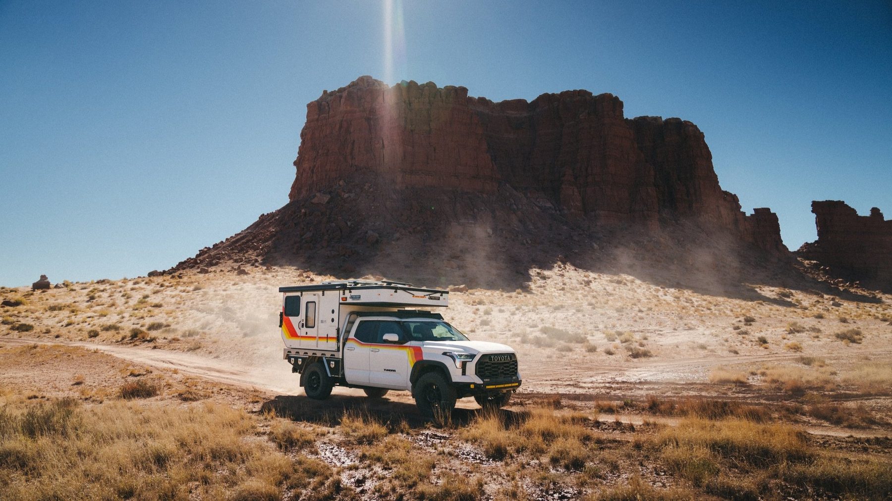 The Tundra in profile, camper roof down, driving along a desert track below a red butte and raising dust