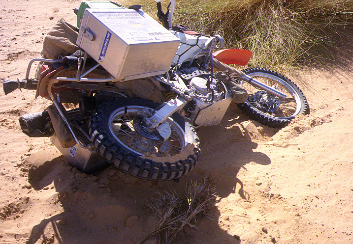 A loaded XR650L lying on its side in soft sand, its aluminium case and a jerrican uppermost, the front wheel half buried