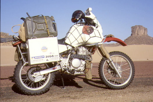 A fully loaded Honda XR650L on its side stand in the desert, with a netted white tank, an aluminium case on the rear rack and a green jerrican strapped on top of the case