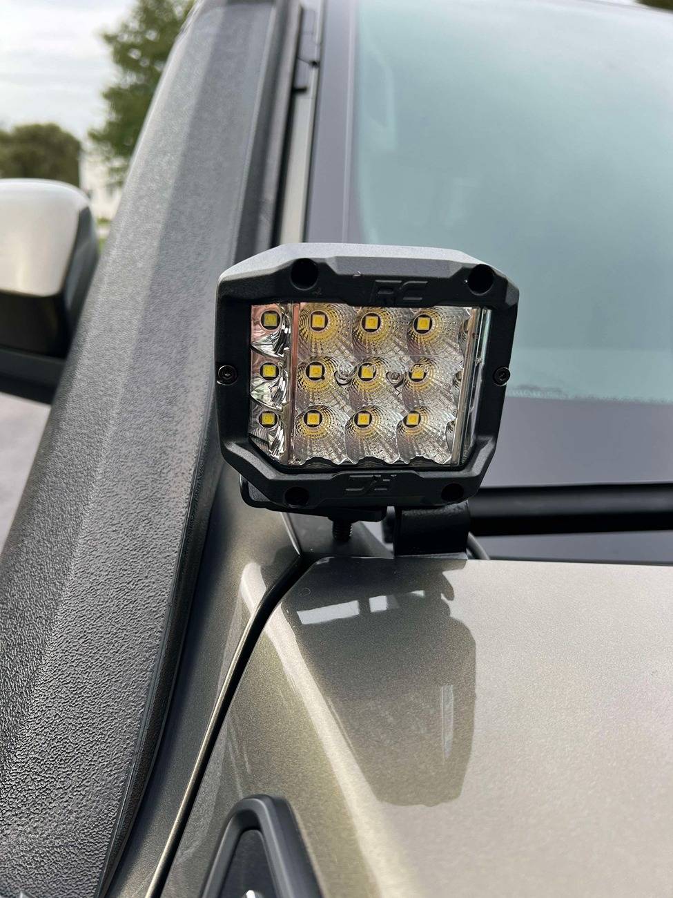 A square LED pod bolted to a bracket at the hood hinge of a Toyota Tacoma, level with the base of the A-pillar and just below the wing mirror