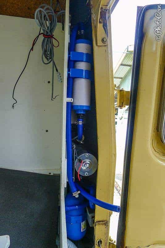 A narrow vertical space in the rear corner of the truck's body, with blue hose, a cylindrical filter and a small pump stacked one above another