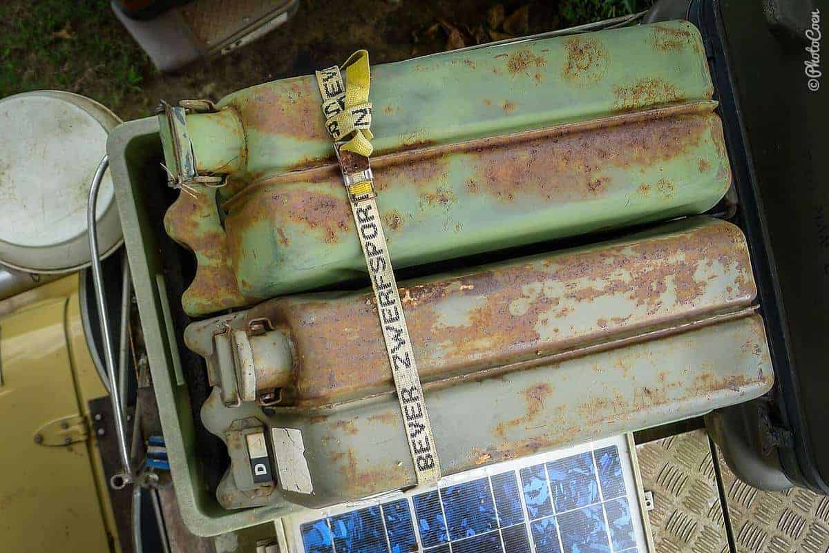 Two rusty green steel jerrycans strapped into a tray on a roof rack beside a small solar panel