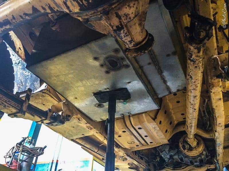 Looking up at the underside of a Land Cruiser in a workshop, a flat steel tank with a round flange held up under the floor by a jack beside the propshaft