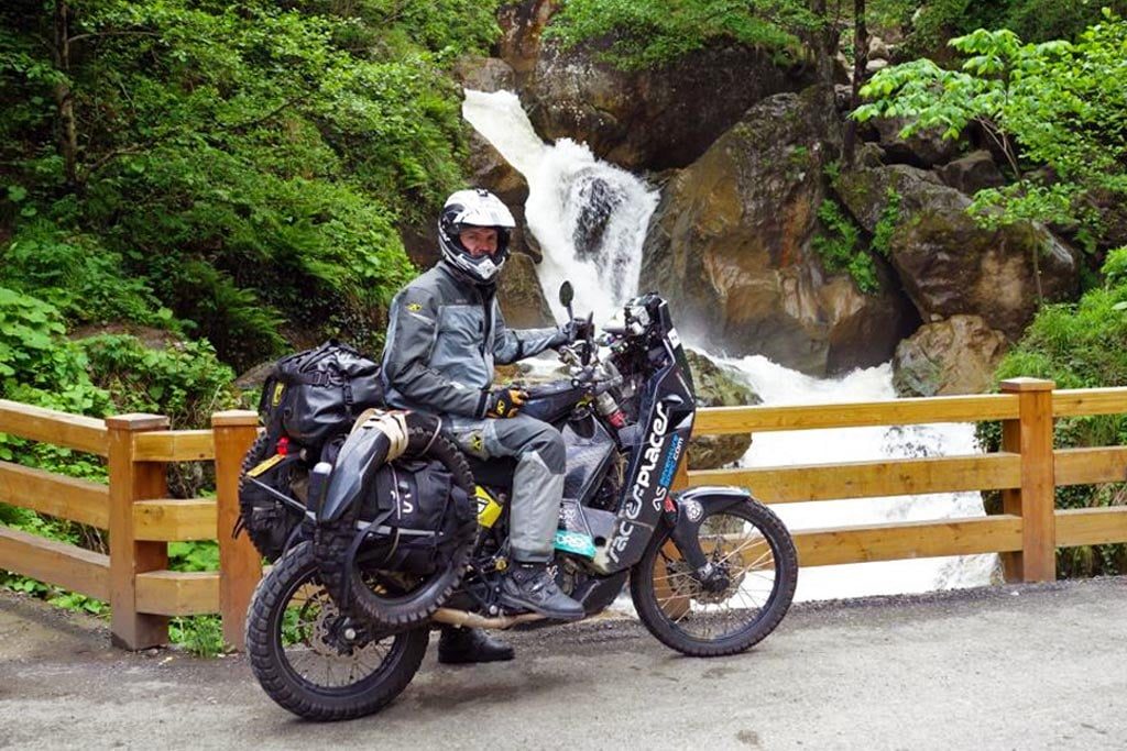 Lyndon Poskitt seated on his loaded KTM 690 Rally beside a waterfall, with panniers, stacked bags and a spare tyre strapped at the back of the bike