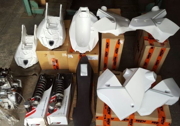 Unpainted white KTM rally bike fuel tanks and bodywork laid out on boxes in a workshop: two long rear tanks and several pairs of side tanks, beside two rear shocks