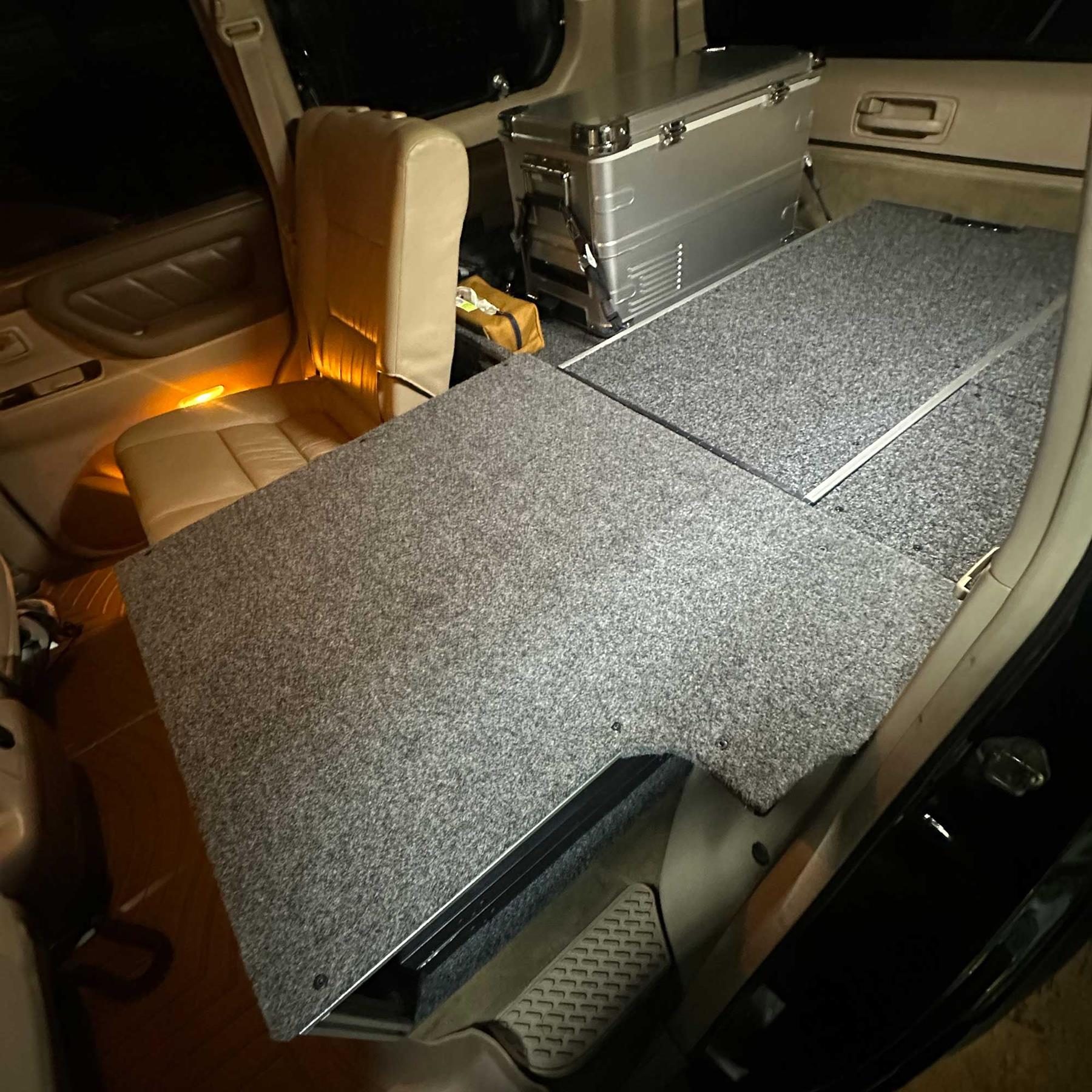 The interior of the Land Cruiser at night: a flat carpeted platform where the second row used to be, a metal fridge behind it, and a single tan leather seat section retained alongside