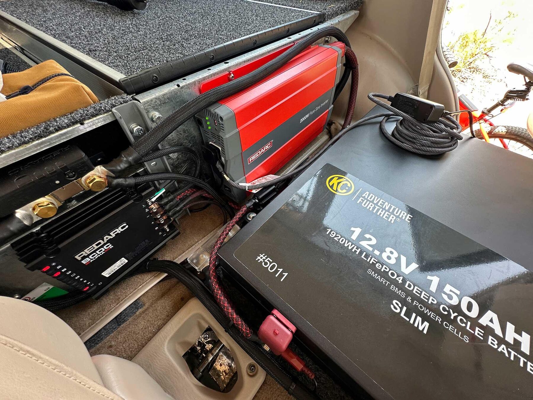 Under a carpeted platform in the back of a Land Cruiser, a slim black KC 12.8 volt 150Ah lithium battery beside a black REDARC DC-DC charger and a red REDARC inverter, with heavy red and black cables between them