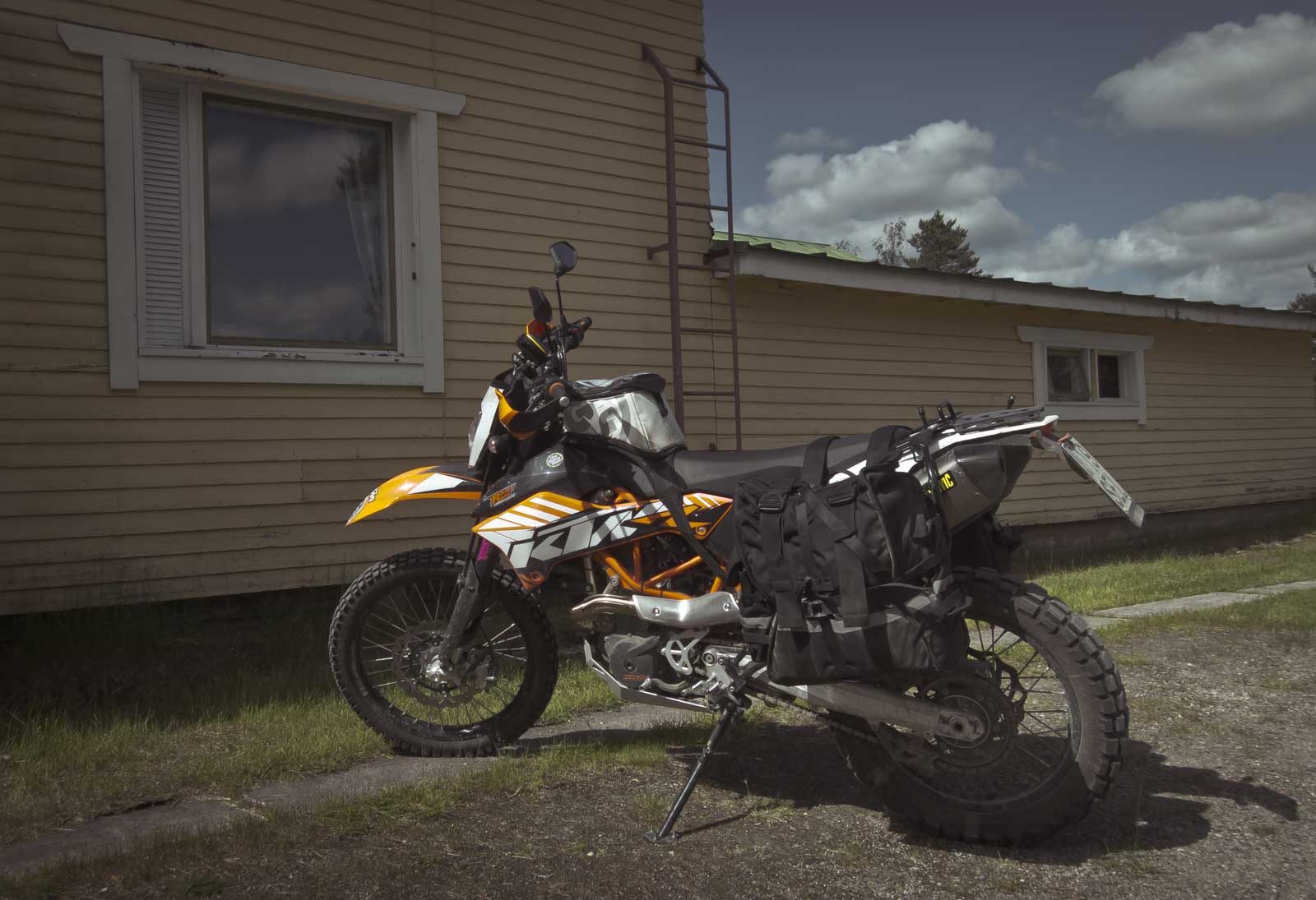 The same KTM 690 earlier in its life, on its side stand beside a wooden house, with black soft panniers hanging low on the left side down near the footpeg and a tank bag on the tank