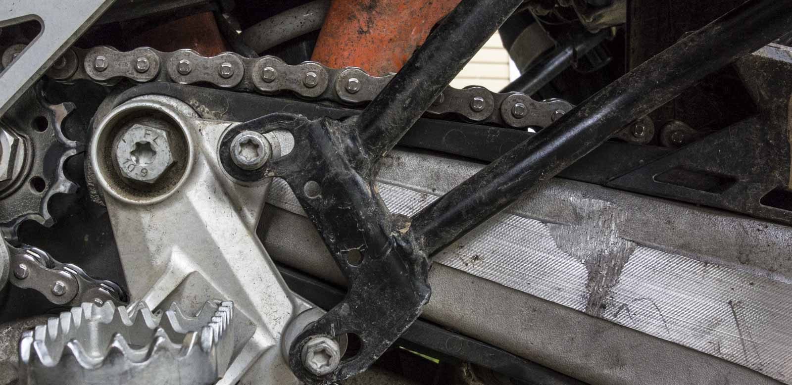 Close-up of the black steel rack's lower leg bolted to the aluminium footpeg bracket, with bright scratches worn into the swingarm beside it