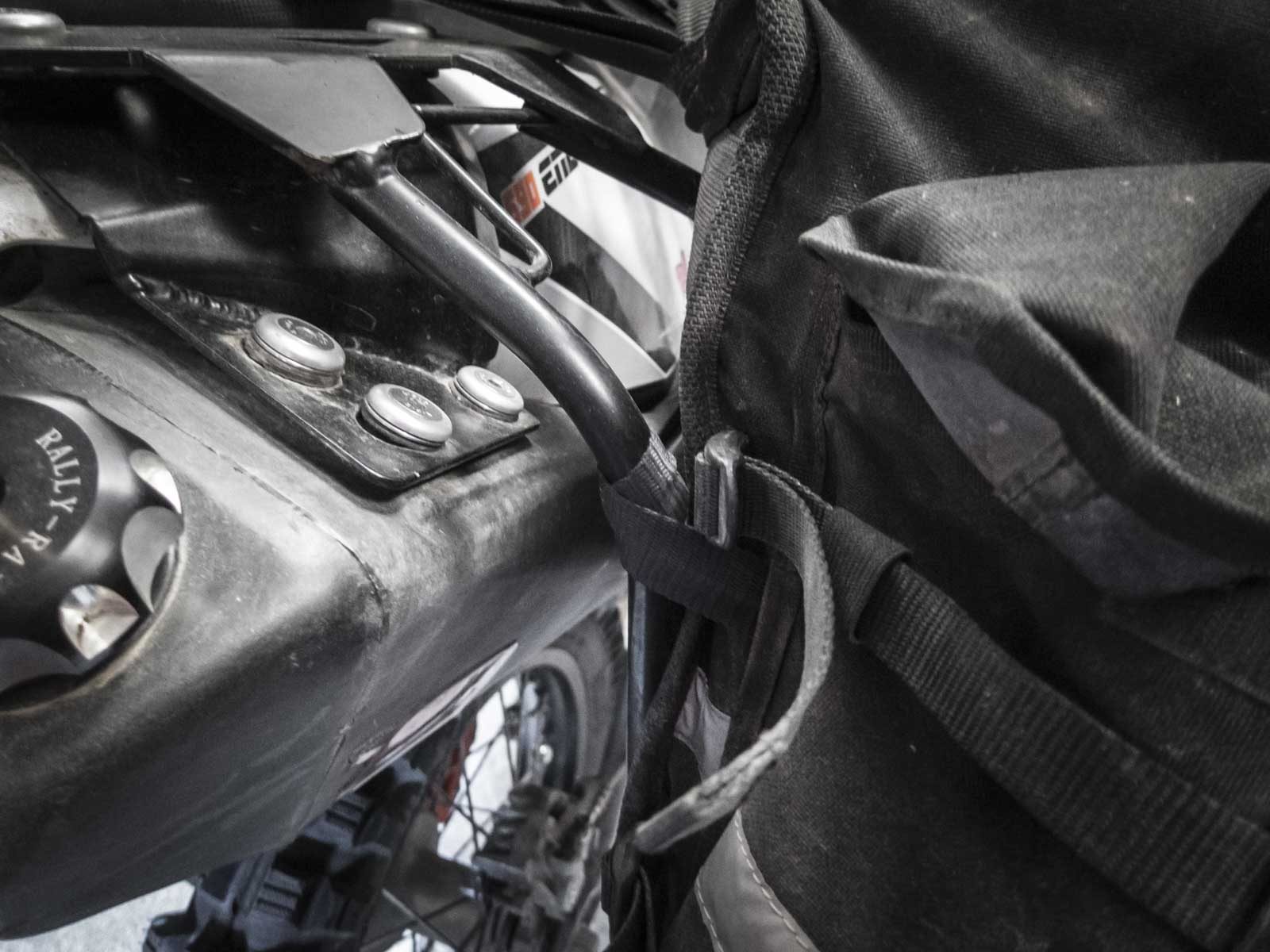 A cargo strap looped round the rack tube beside the tail of the bike, next to a rack plate bolted to the plastic tail with three large bolts