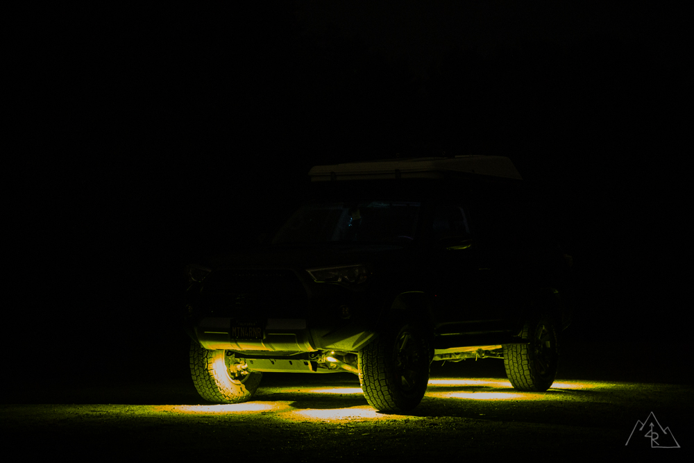 A dark 4Runner at night seen from the front quarter, its body barely visible, with pools of amber light on the grass under and beside the front wheel, under the sliders and out past the tires