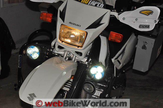 The front of a white Suzuki DR650 in a garage at night, with its yellowish halogen headlight on and two small round LED lights glowing bright white either side of the front fender